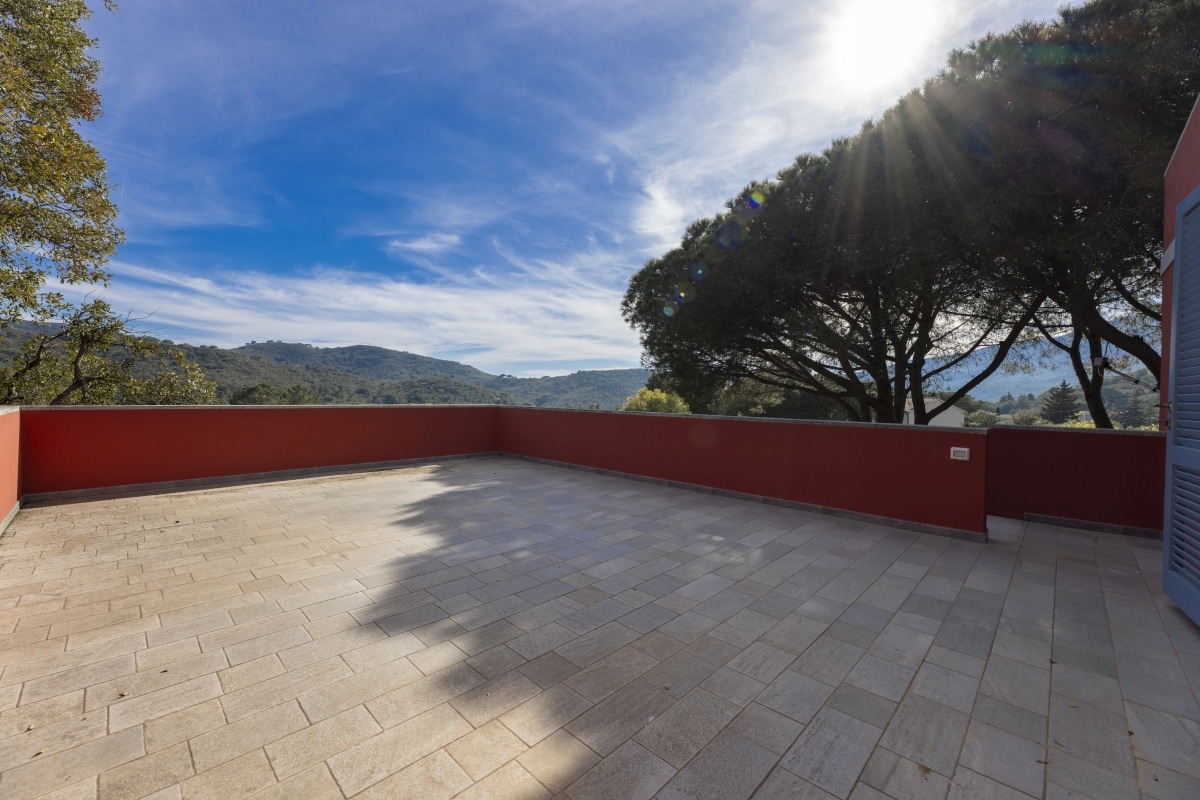 A spacious terrace is revealed, featuring stone flooring and surrounded by lush greenery. The view encompasses rolling hills under a bright sky, with sunlight casting gentle rays across the open space. This area allows for relaxation and enjoyment of the natural surroundings.