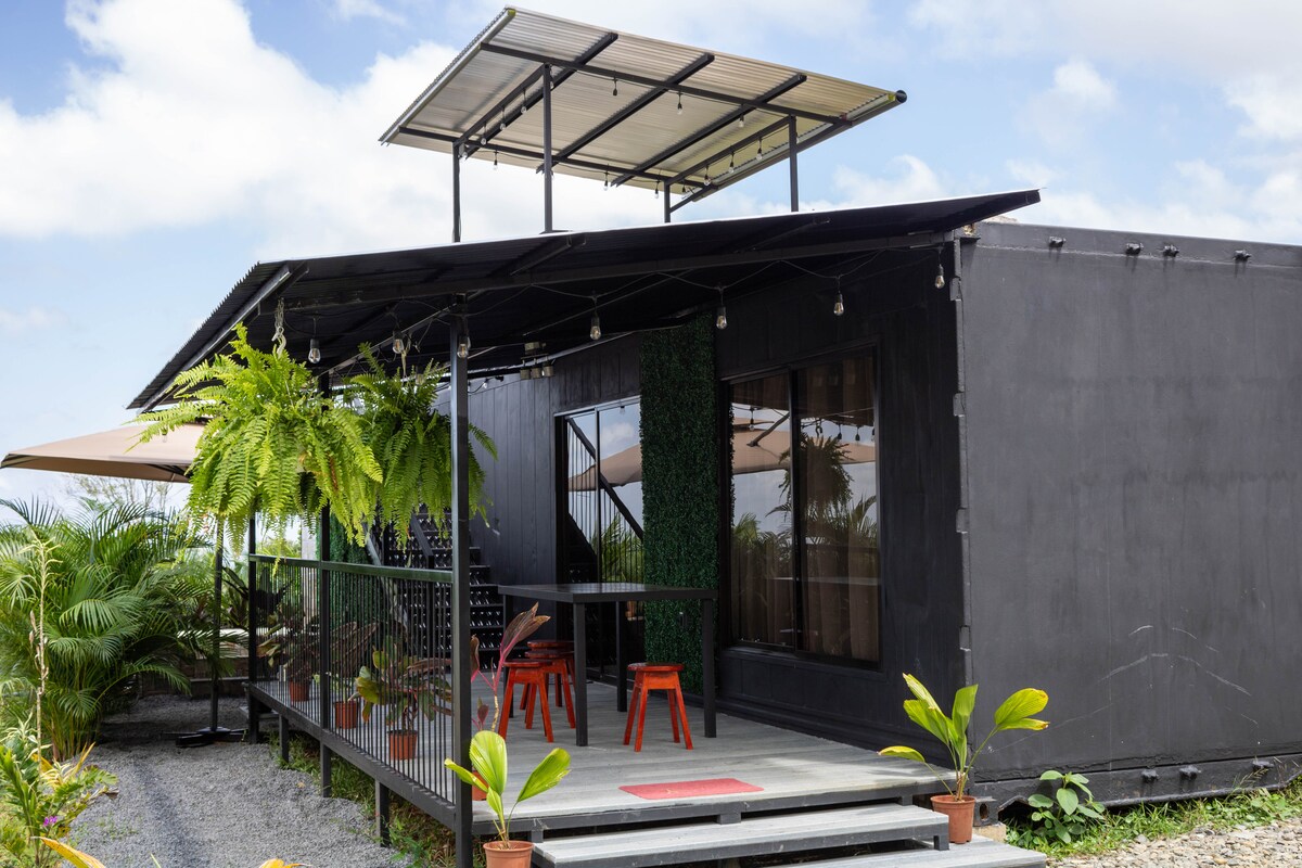 A modern structure is surrounded by lush greenery, featuring a spacious patio with seating and a stylish roof overhang. Large windows reflect natural light, while potted plants add a touch of nature to the entrance area.