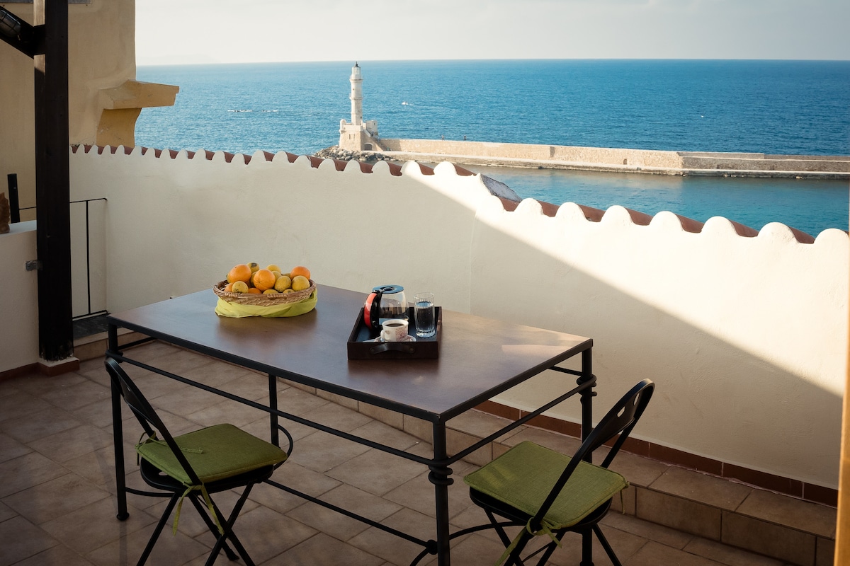 A roof terrace with a table set for breakfast, featuring a bowl of fresh oranges and coffee cups. Two green chairs are positioned at the table, with the view overlooking the serene blue sea and the coastal Lighthouse in the distance.