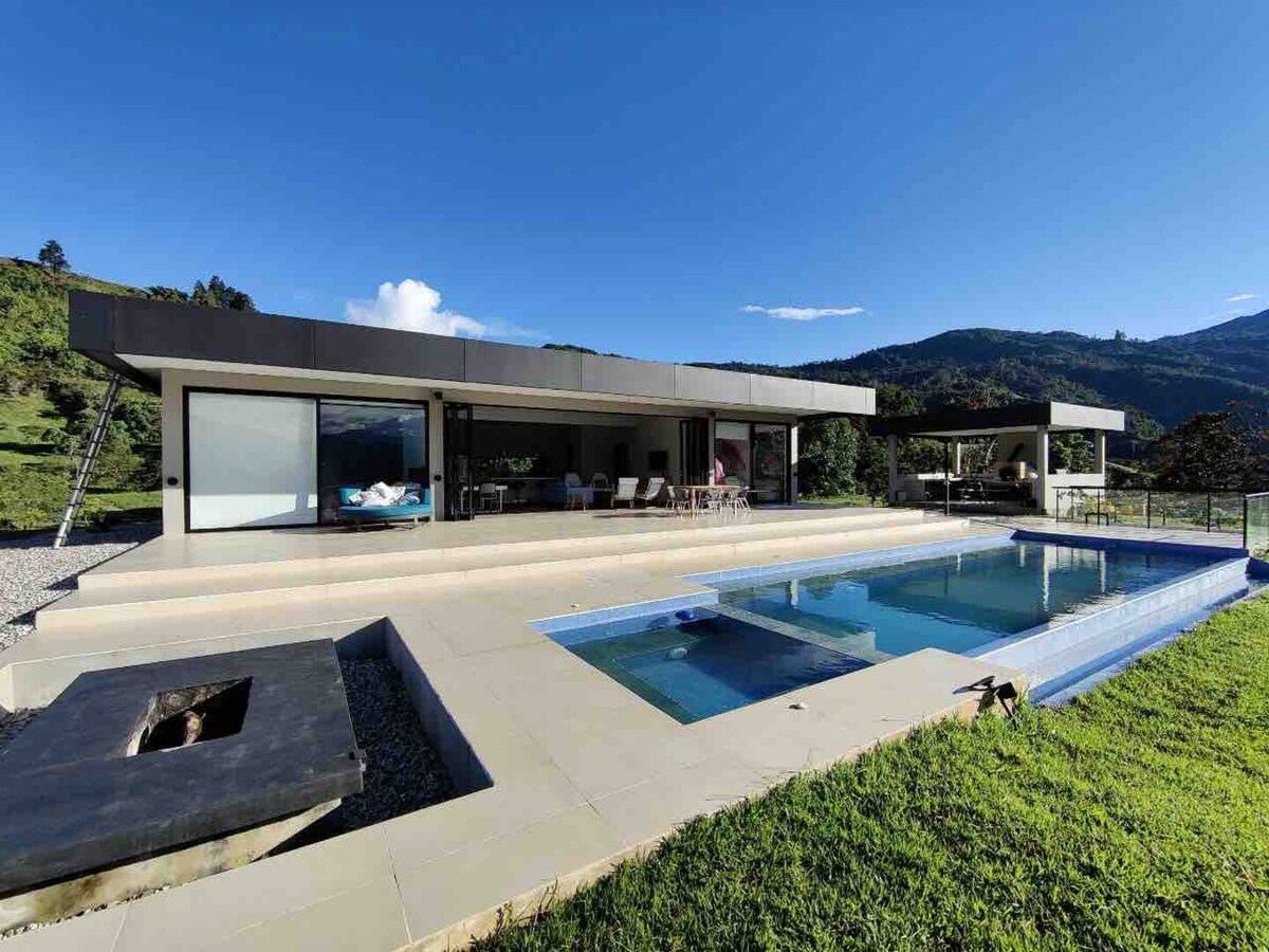An expansive outdoor area features a modern pool reflecting the surrounding mountains. The structure includes large glass walls that allow for views of the serene natural landscape. Comfortable seating is arranged on a patio, inviting relaxation amid the tranquil setting.