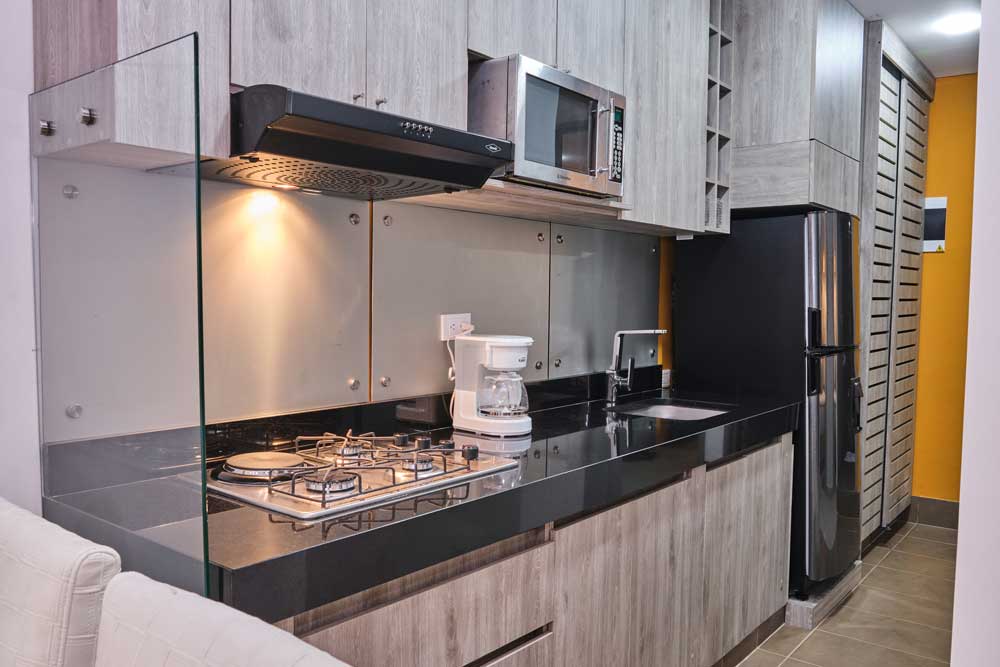 A modern kitchen features sleek wooden cabinetry and a black countertop. Stainless steel appliances, including a microwave and coffee maker, are visible, along with a four-burner gas stove. Warm lighting enhances the organized layout, contributing to a functional cooking space.