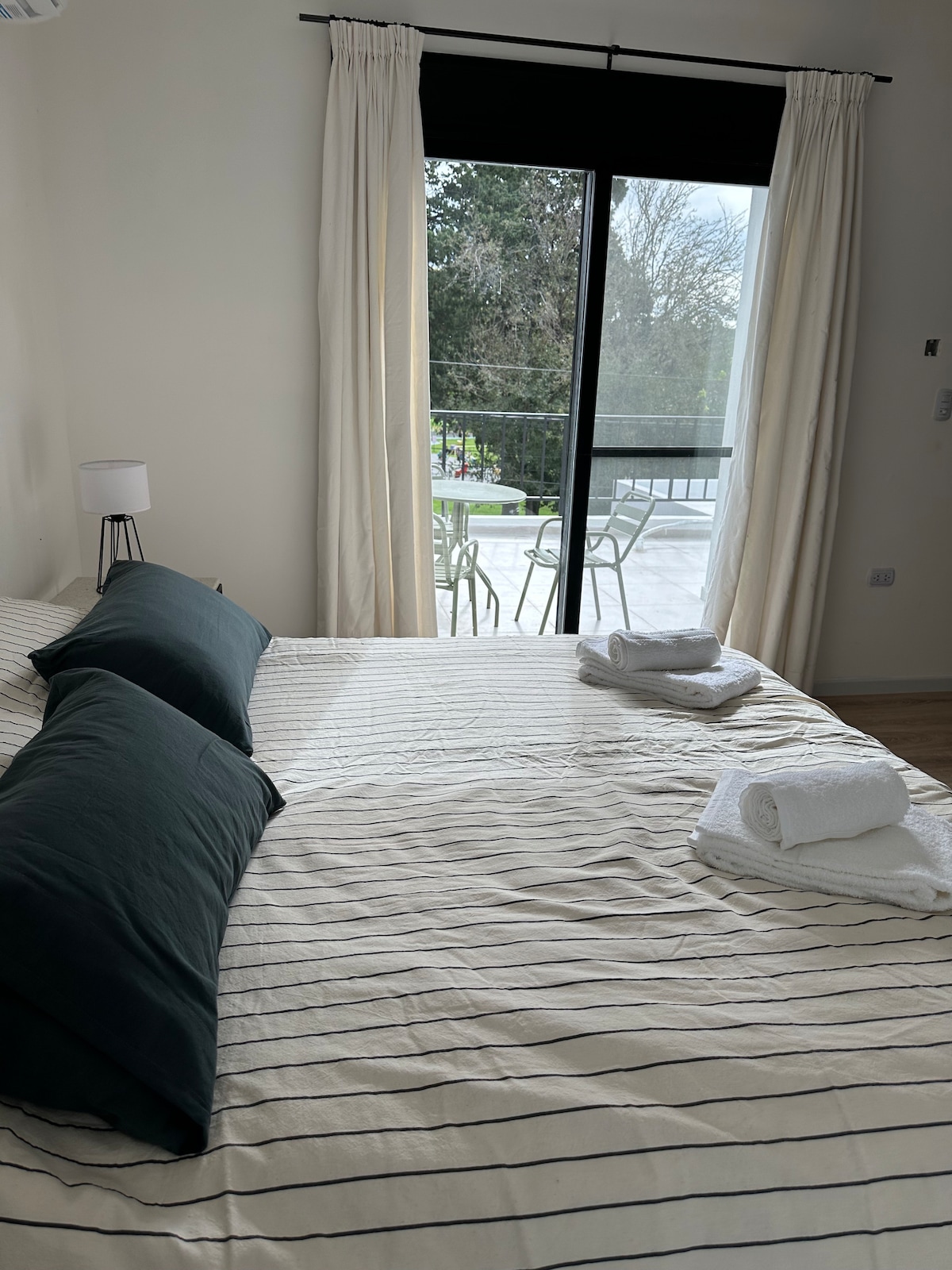 A spacious sleeping area is highlighted by a neatly made bed with striped linens and two dark green pillows. Bright light filters through sliding glass doors, revealing a private terrace with outdoor furniture and a view of the greenery outside.