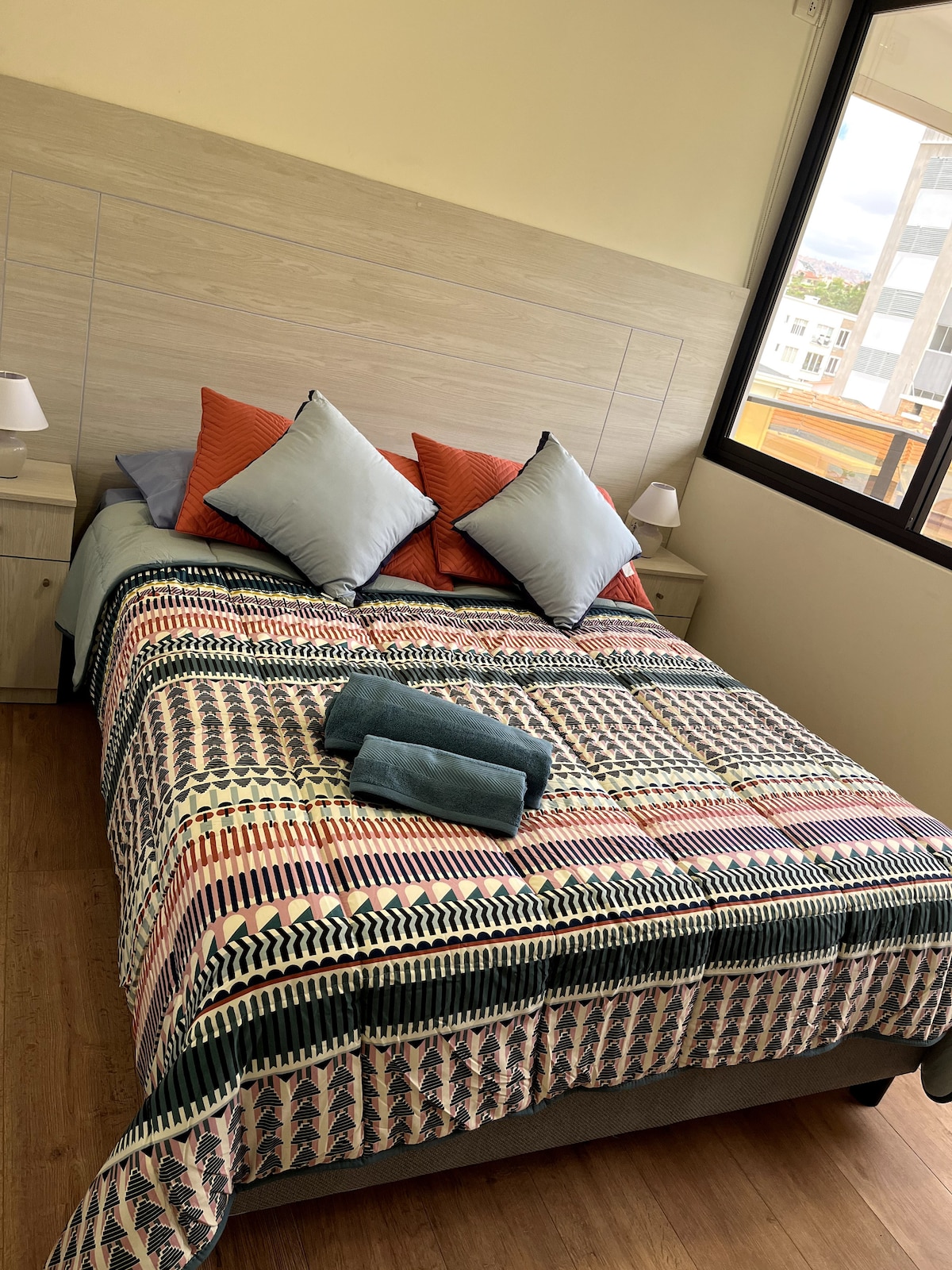 A well-appointed bedroom is featured with a patterned bedspread over a spacious bed. Two pillows with contrasting colors are positioned on top of the bed. Nightstands with lamps are located on either side, and large windows allow natural light to fill the space.