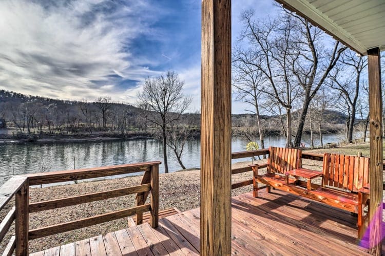 A serene view of a river is framed by wooden beams. Two rustic wooden chairs are positioned on the deck, facing the water. Leafless trees line the bank, while the distant hills are gently outlined against a blue sky with wispy clouds.