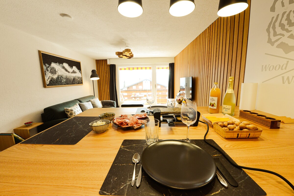 A dining setup is prominently displayed on a textured wooden table, featuring elegant glassware and black plates. In the background, a cozy living area with a dark sofa is visible, complemented by a scenic view of mountains through large windows.