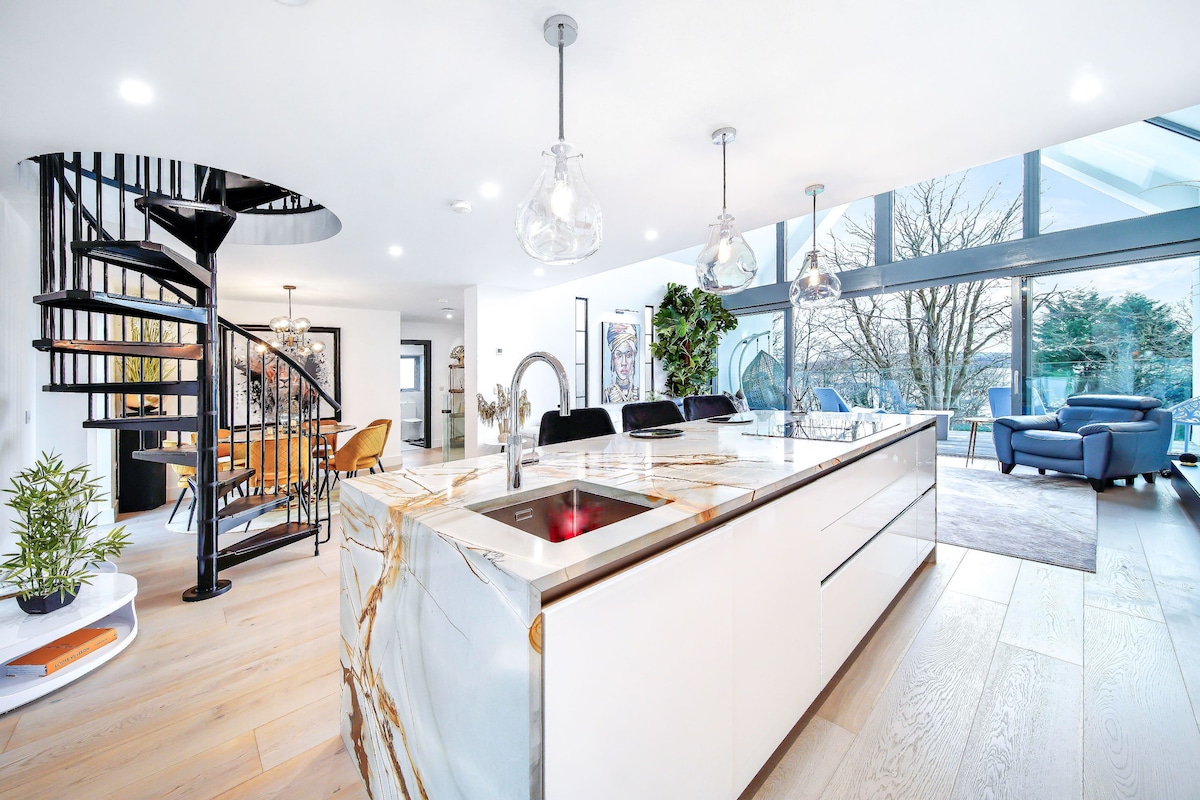 An open plan kitchen showcases sleek cabinetry with a striking patterned countertop. Modern pendant lights hang above the central island, which features a built-in sink. A spiral staircase leads to the upper floor, while large glass doors offer views of the surrounding nature.