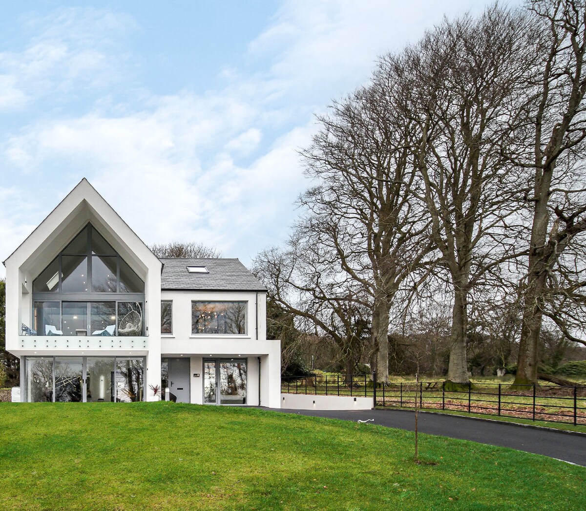 The modern structure features a distinctive triangular roof and large glass windows, allowing natural light to fill the interior. A lush green lawn surrounds the home, while tall trees provide a serene backdrop, creating a tranquil setting that blends nature and contemporary design.