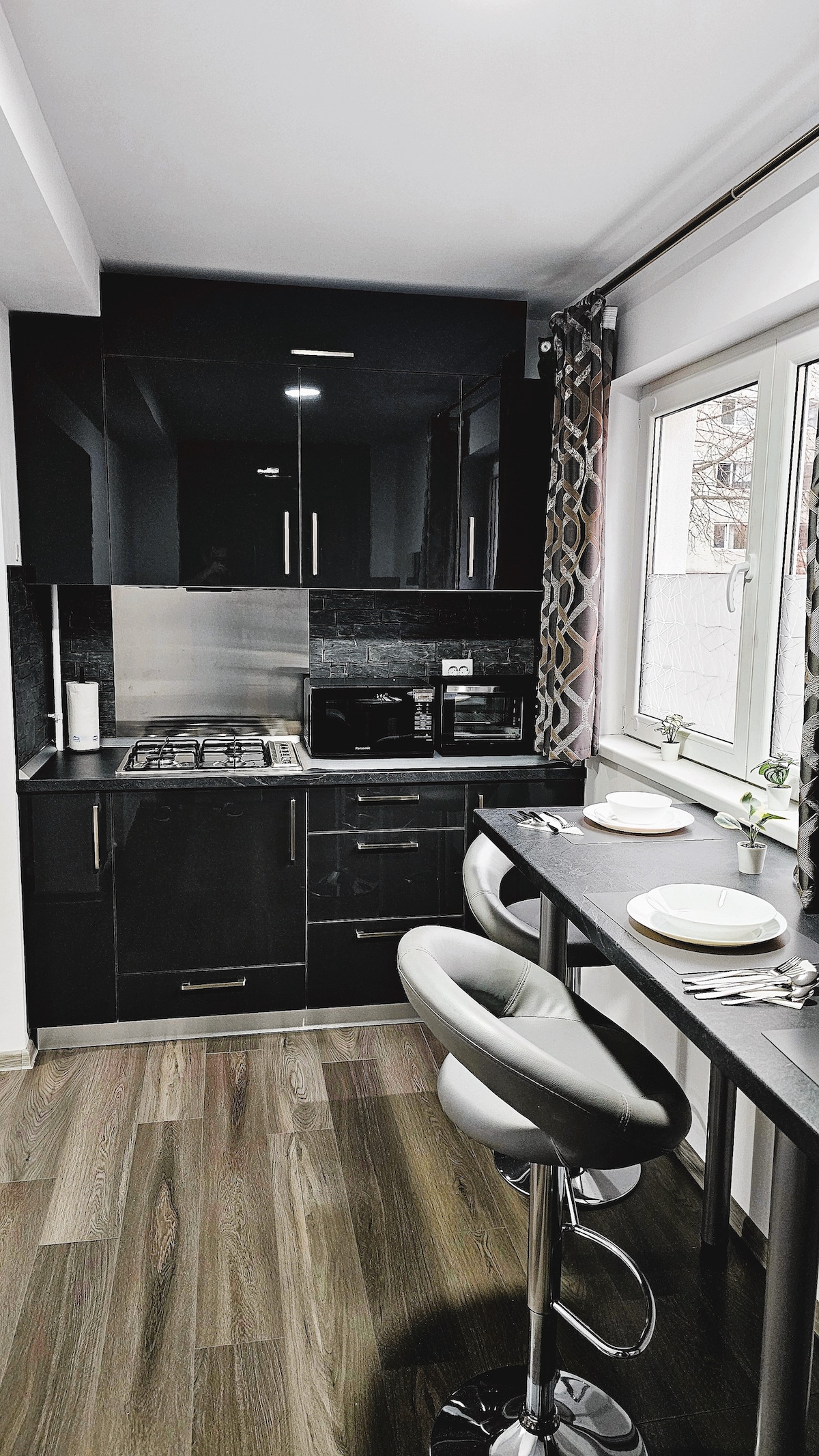 A modern kitchen is presented, featuring sleek black cabinetry and stainless steel appliances. A small dining area is visible with two bar stools and a compact table set for two. Natural light enters through a window, enhancing the kitchen's contemporary design.