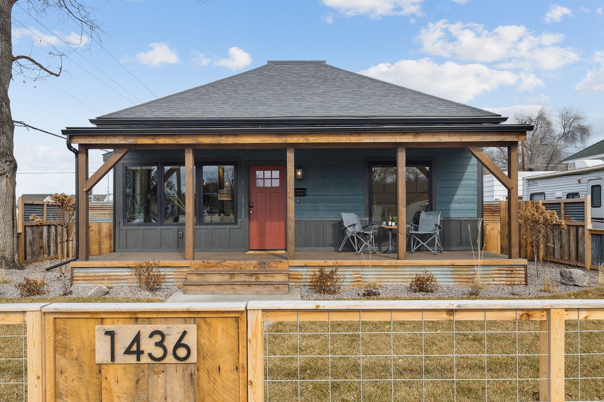 A charming mountain modern home is presented with a welcoming red front door. A spacious porch features two grey chairs, framed by wooden beams. Gravel landscaping surrounds the entrance, while a wooden fence adds a rustic touch, enhancing the property’s appeal.