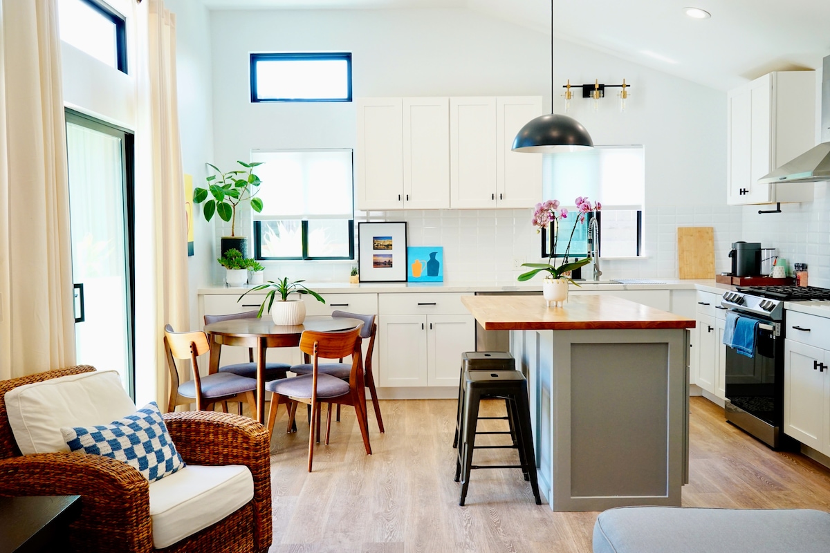 An open concept kitchen and dining area features white cabinetry and modern appliances. A wooden island provides seating, while a round dining table is set for four. Natural light floods the space through large windows, complemented by greenery and decorative touches.