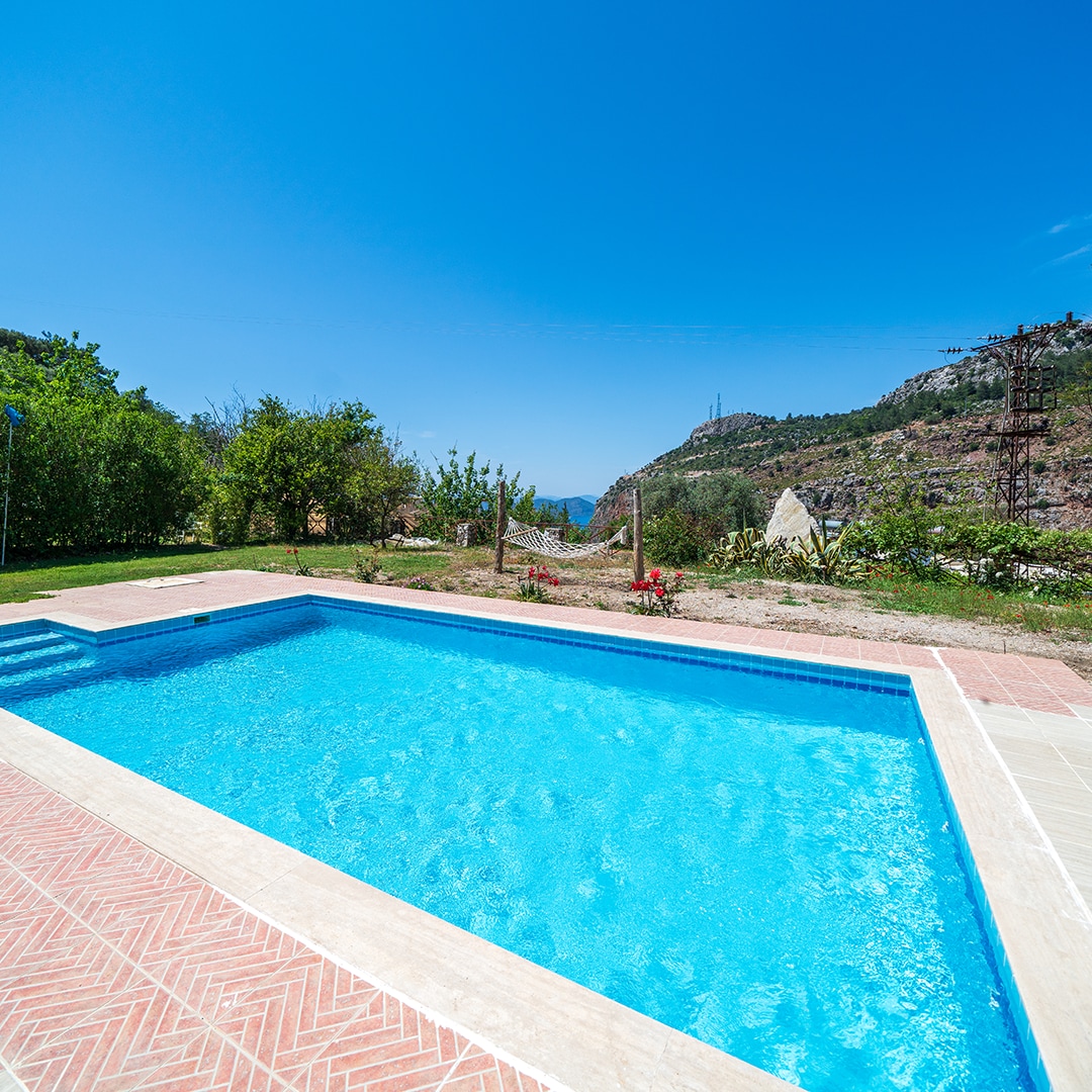 A clear blue pool is surrounded by well-maintained greenery, set against a backdrop of rolling hills. The water sparkles under the sunlight, and hints of colorful flowers emerge from the surrounding landscape.