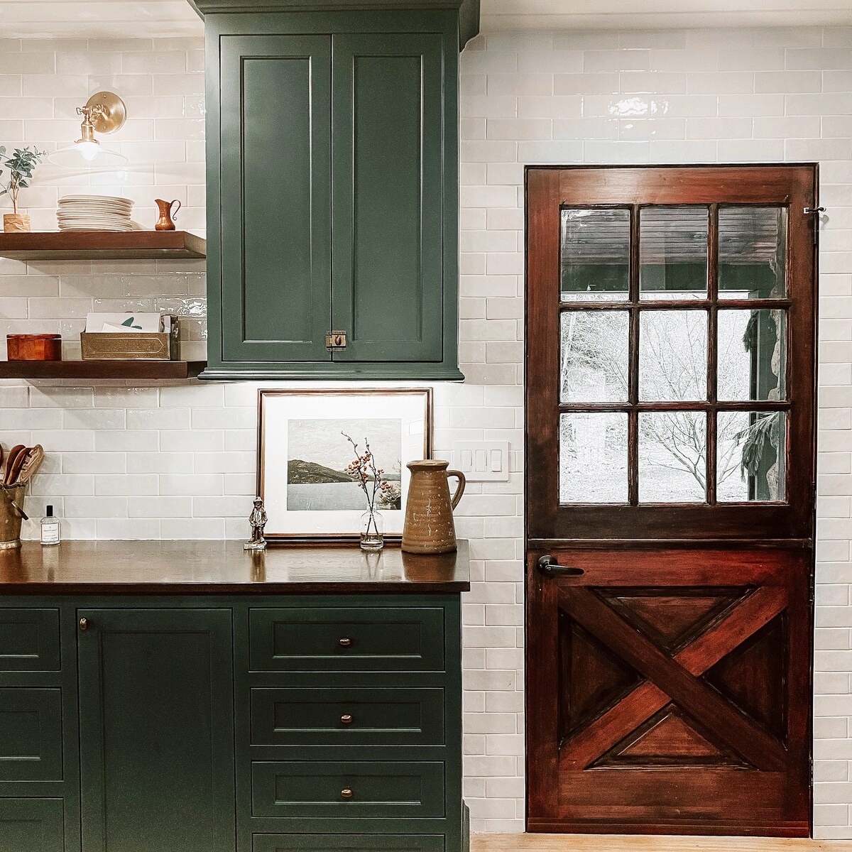 The kitchen features deep green cabinetry with gold accents, showcasing neatly arranged dishware on open shelves. A rustic wooden door with glass panes complements the space, while a framed artwork adds a touch of charm. Natural light brightens the area, enhancing the inviting atmosphere.