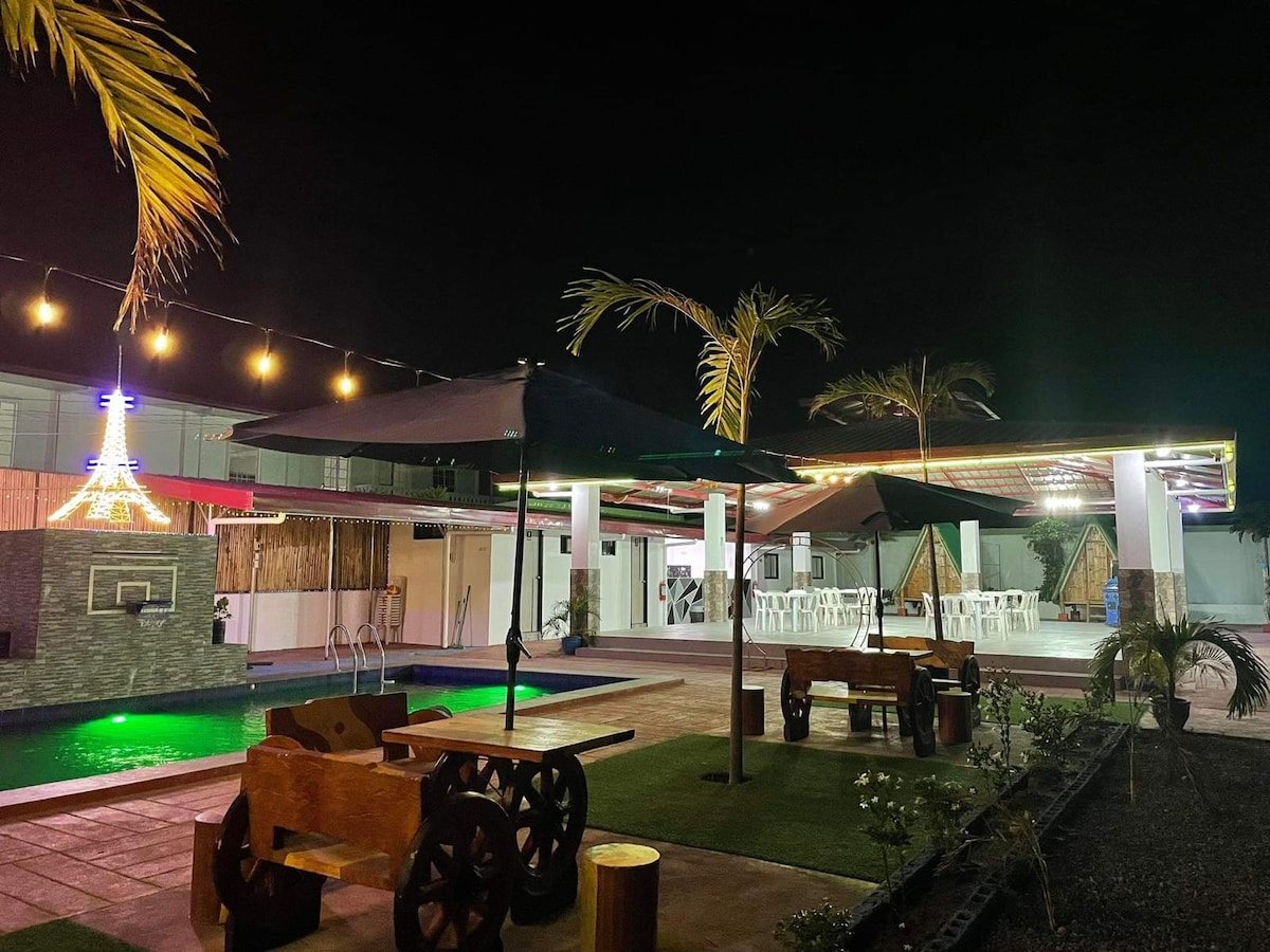 An outdoor recreational area is seen at night, enhanced by decorative lighting. Wooden tables and chairs are arranged on the patio, while palm trees provide a tropical feel. The space includes a pool and shaded areas, fostering relaxation and social gatherings.