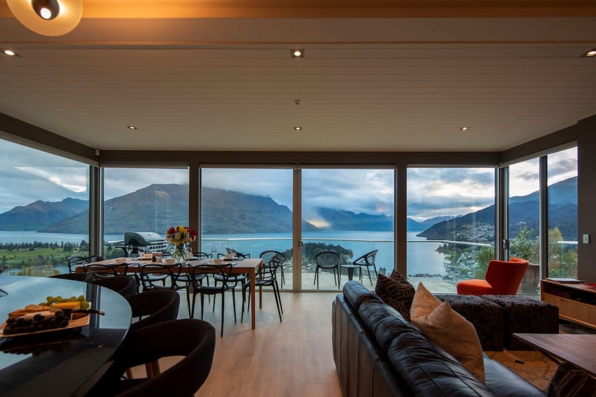 An open living area is framed by large windows offering expansive lake and mountain views. A dining table is set with chairs nearby, while a cozy seating arrangement features a dark couch and a bright orange chair. Natural light fills the space, creating a serene ambiance.