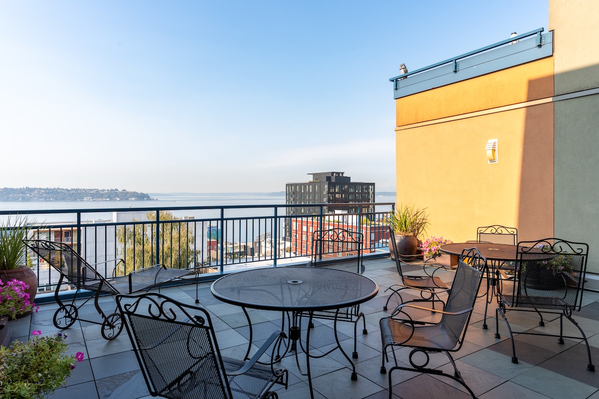 A rooftop terrace showcases multiple seating arrangements with metal tables and chairs. Lush planters with greenery and flowers are positioned along the railing. A clear view of Elliott Bay and the surrounding area enhances the outdoor space, complemented by a bright and open sky.