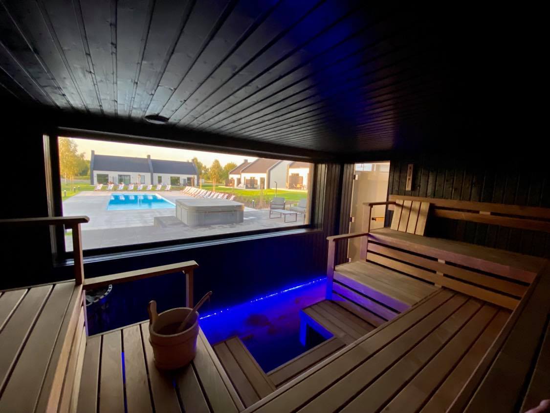 A wooden sauna interior features benches arranged around a small, circular opening. Soft blue lighting highlights the seating area, while a large window provides a view of the outdoor relaxation space, including a heated pool and jacuzzi.