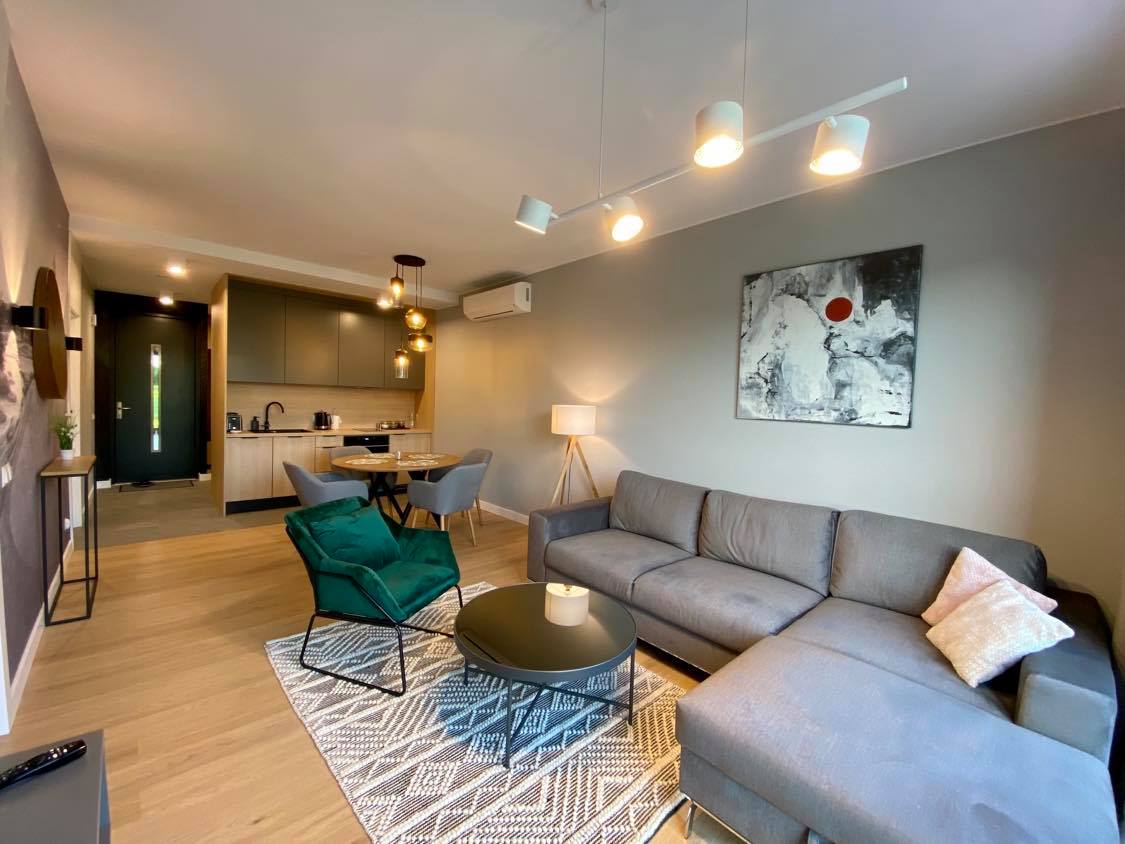 A modern living area features a sectional sofa in neutral tones and a round coffee table. An area rug adds texture to the wood flooring. The space is illuminated by pendant lights, and the open kitchen can be seen in the background, equipped with contemporary appliances.