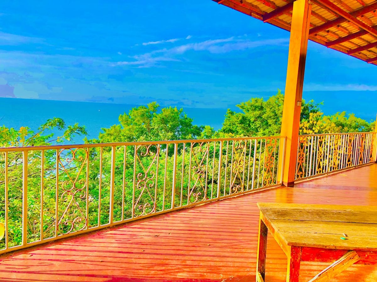 A spacious wooden deck features a railing adorned with decorative patterns, looking out over lush greenery and the expansive view of Lake Victoria. The bright blue sky complements the colorful setting, inviting a serene atmosphere.