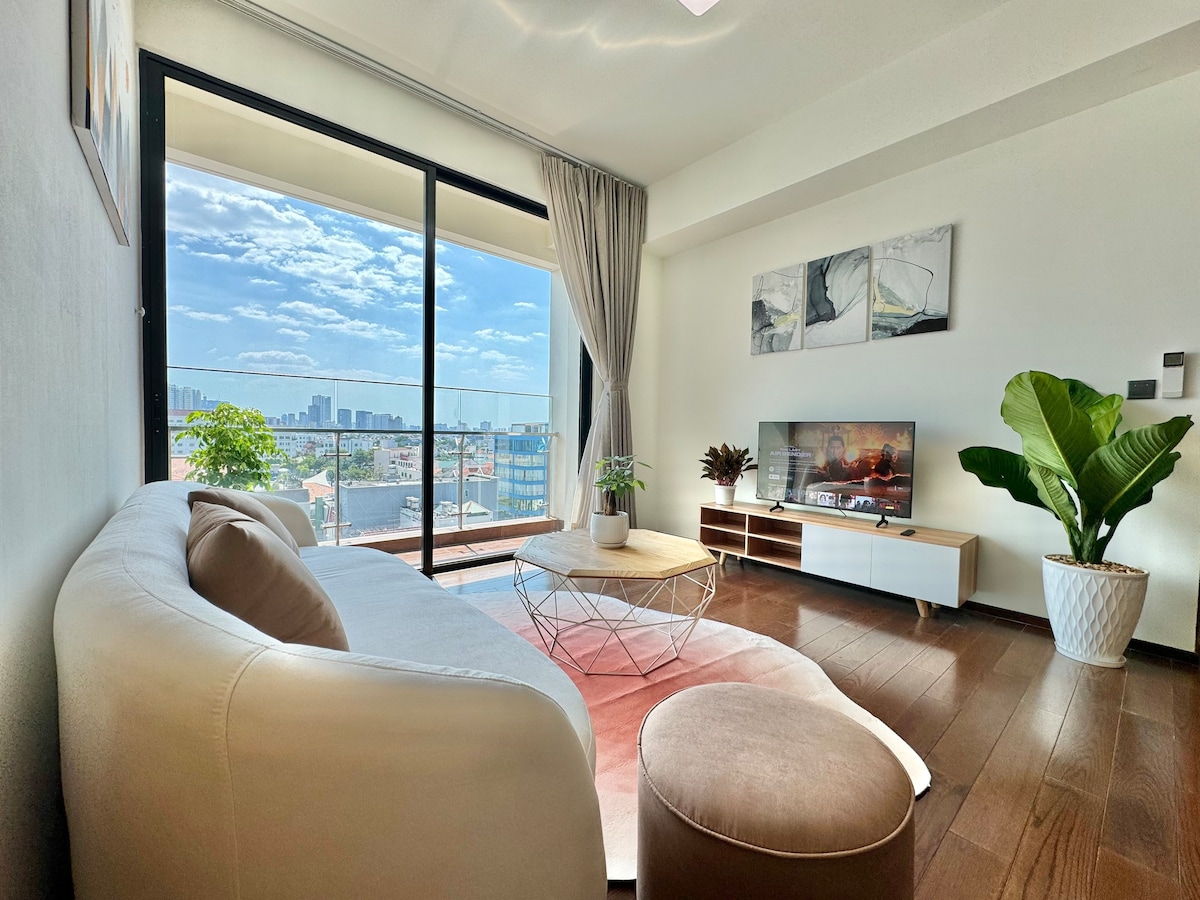 A modern living space is displayed with a spacious white sofa and a circular coffee table. Natural light fills the room through large floor-to-ceiling windows, showcasing a city view. A green plant and decorative items enhance the welcoming ambiance.
