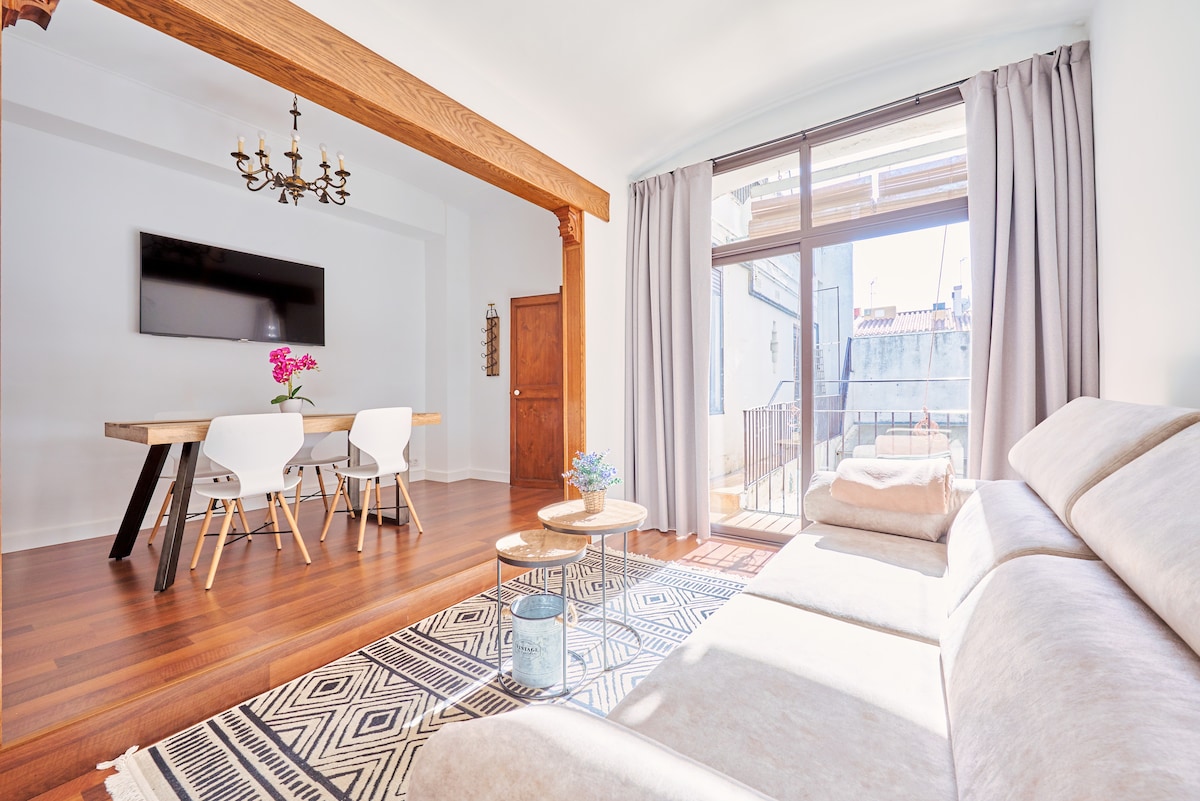 A spacious living area is highlighted by minimalist décor, featuring a large sectional sofa and a contemporary dining table with white chairs. Natural light filters through glass doors that open to a balcony, while a potted plant and decorative items adorn the coffee table.