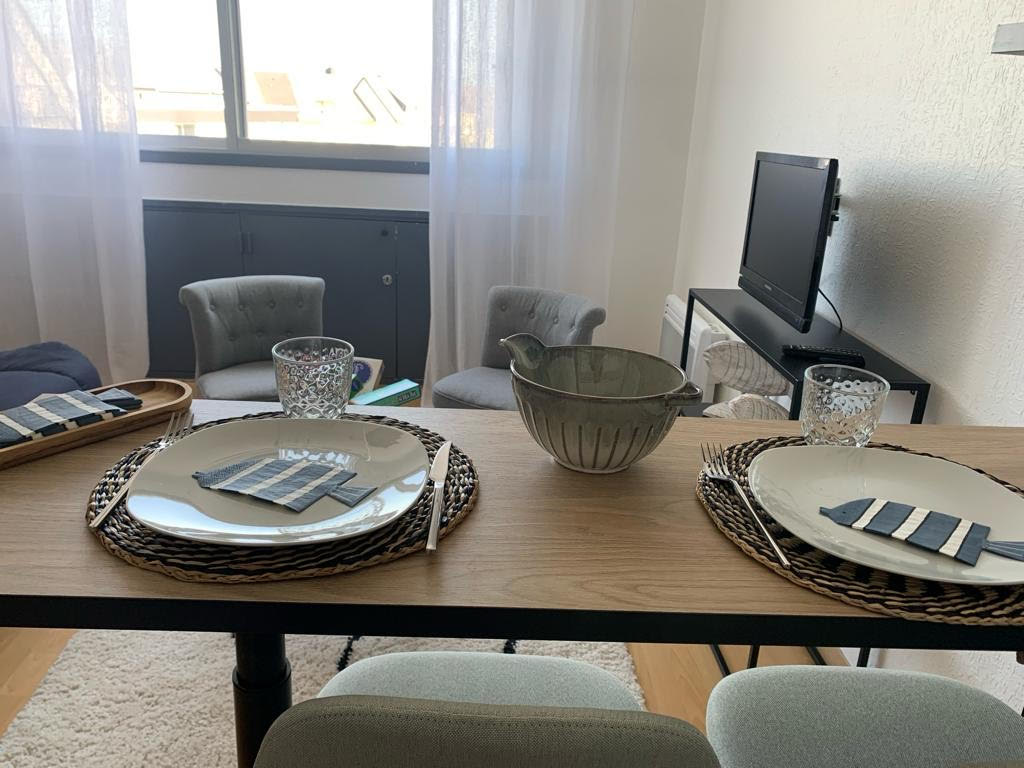 A dining table is set for three, featuring decorative plates and woven placemats. Glassware adds a touch of elegance, while a decorative bowl sits in the center. Light filters through sheer curtains, illuminating the space with a soft glow.