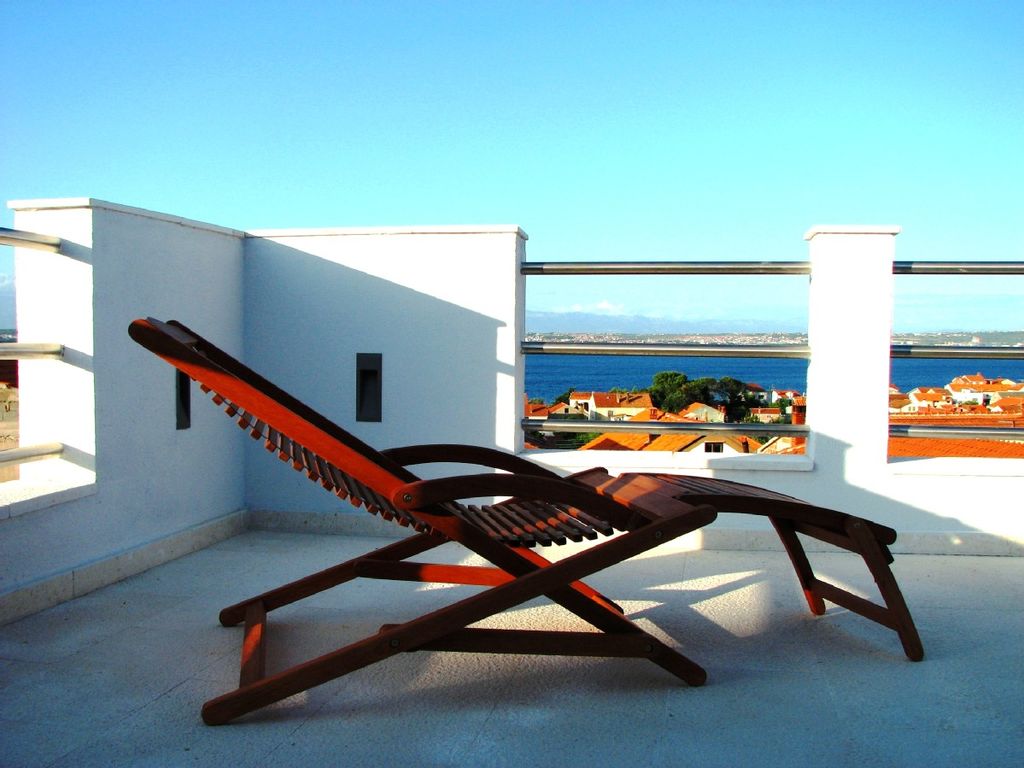 A wooden lounge chair is positioned on a private terrace, providing expansive views of the blue sea and distant land. The clear sky enhances the serene atmosphere, while the surrounding architecture features white walls and a railing.