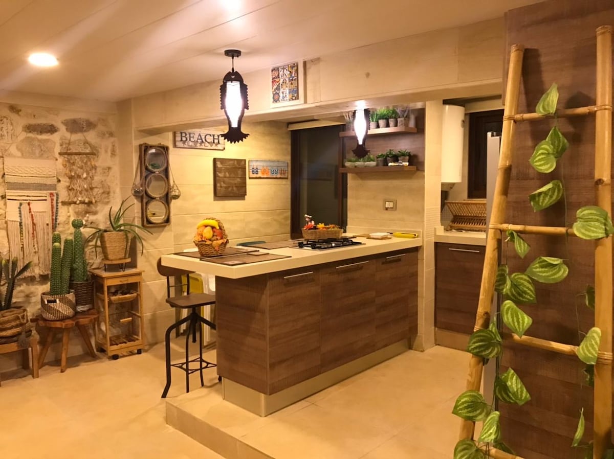 A modern kitchen area showcases earthy tones and natural textures. Wooden cabinetry complements beige stone walls. A breakfast bar with high stools provides seating, while potted plants and decorative items enhance the inviting ambiance. A bamboo ladder leans decoratively against the wall.