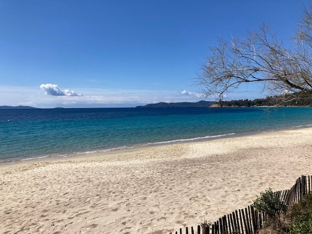 A sandy beach stretches into the distance, bordered by gentle waves in varying shades of blue. A clear sky overhead is framed by a few wispy clouds, and a dune grass fence lines the shore, contributing to the serene coastal view.