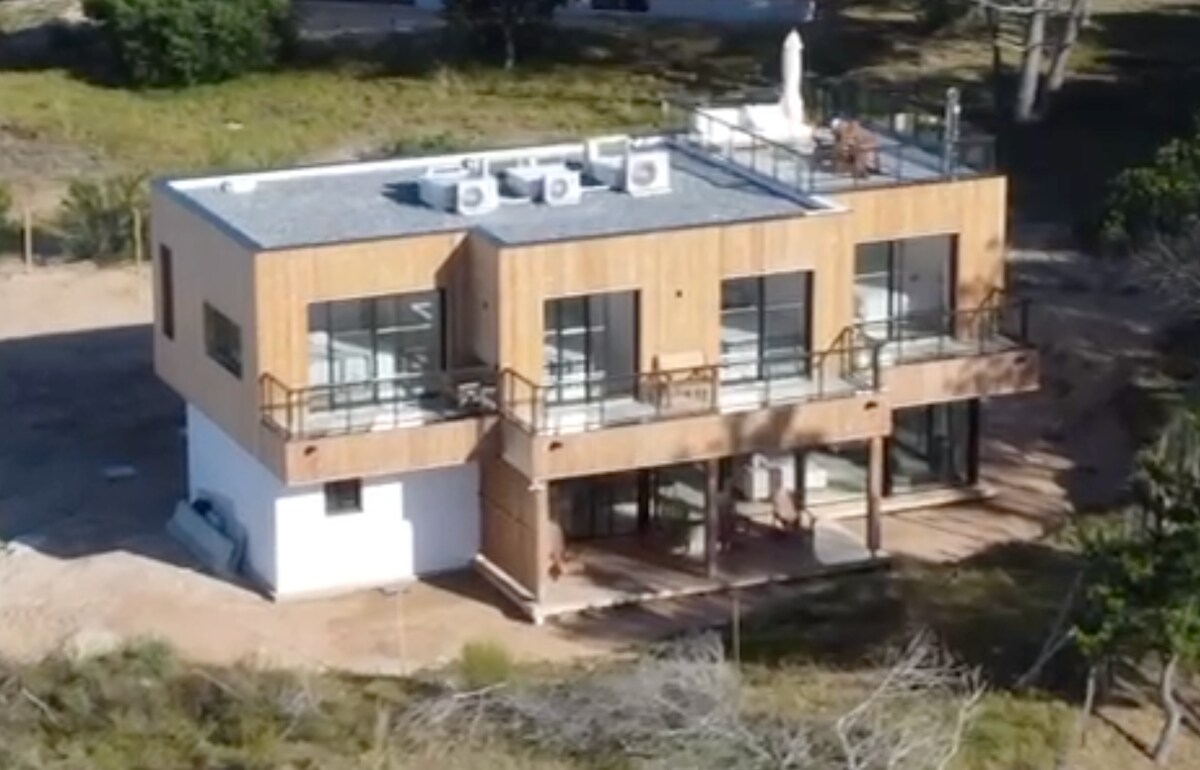 The exterior of the beach house is showcased, featuring a modern design with wooden facades and expansive glass windows. A rooftop terrace is visible, surrounded by greenery and spacious outdoor areas, while multiple air conditioning units are installed on the roof.