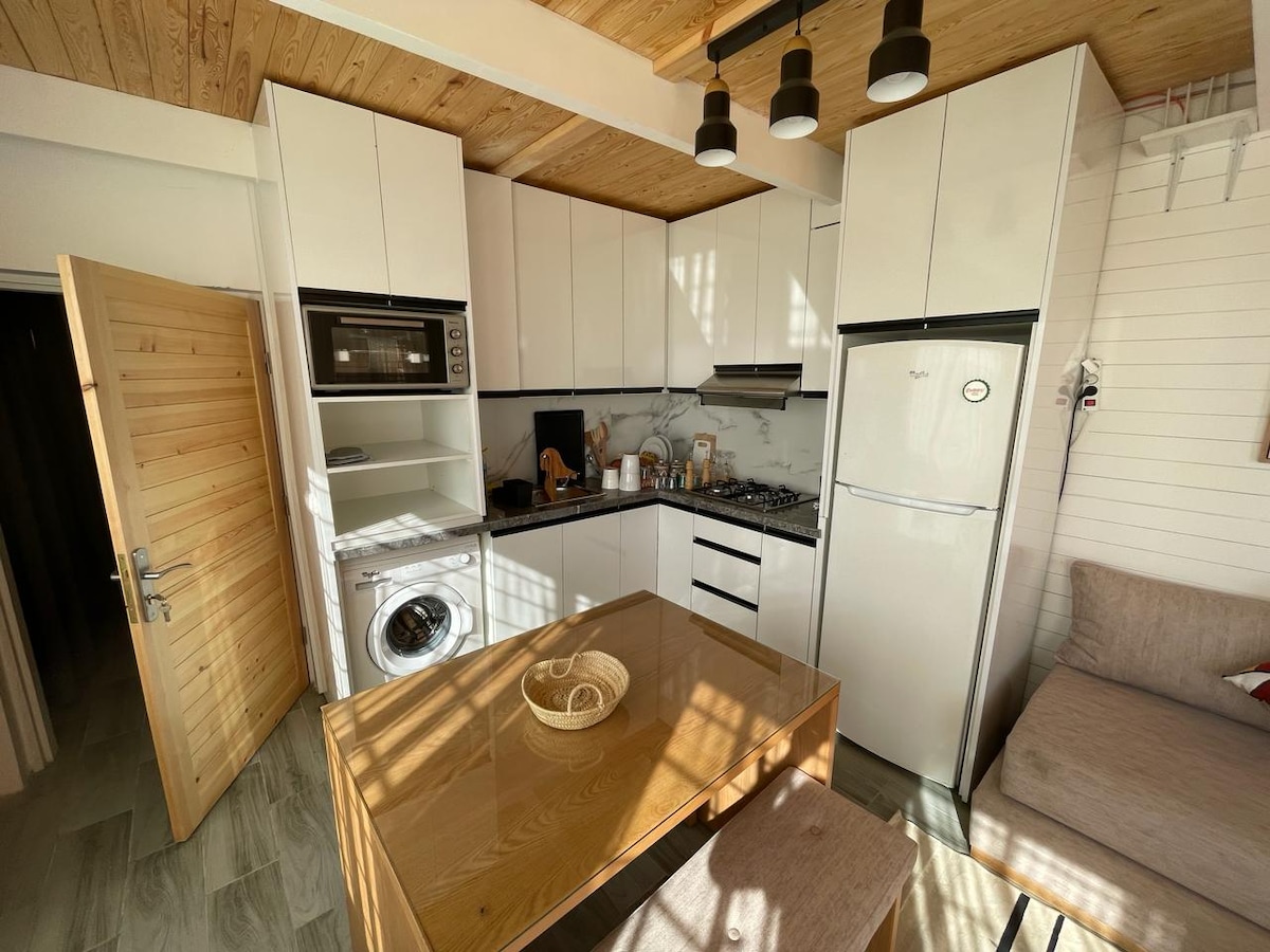 A modern kitchen is equipped with sleek white cabinets, a gas stove, a refrigerator, and a microwave. An adjacent dining table sits beneath a series of pendant lights. Natural light filters through the space, creating a bright and inviting atmosphere.