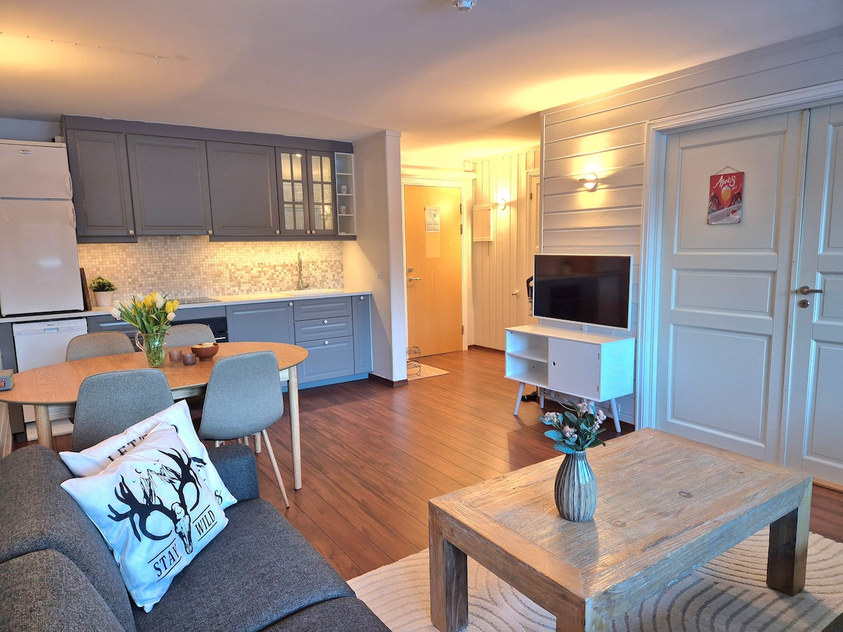 A modern living space features a cozy seating area with a neutral sofa, a wooden coffee table, and a dining table set for four. The kitchen is equipped with gray cabinetry and appliances, while warm lighting enhances the room's welcoming ambiance.