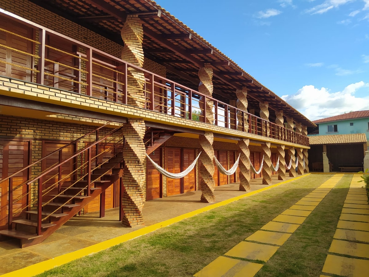 The exterior of a multi-story building features a brick façade with wooden balconies and railings. A stone pathway lined with greenery leads to the entrance. Sturdy staircase structures provide access between the levels, and the sky is clear with soft, natural lighting.