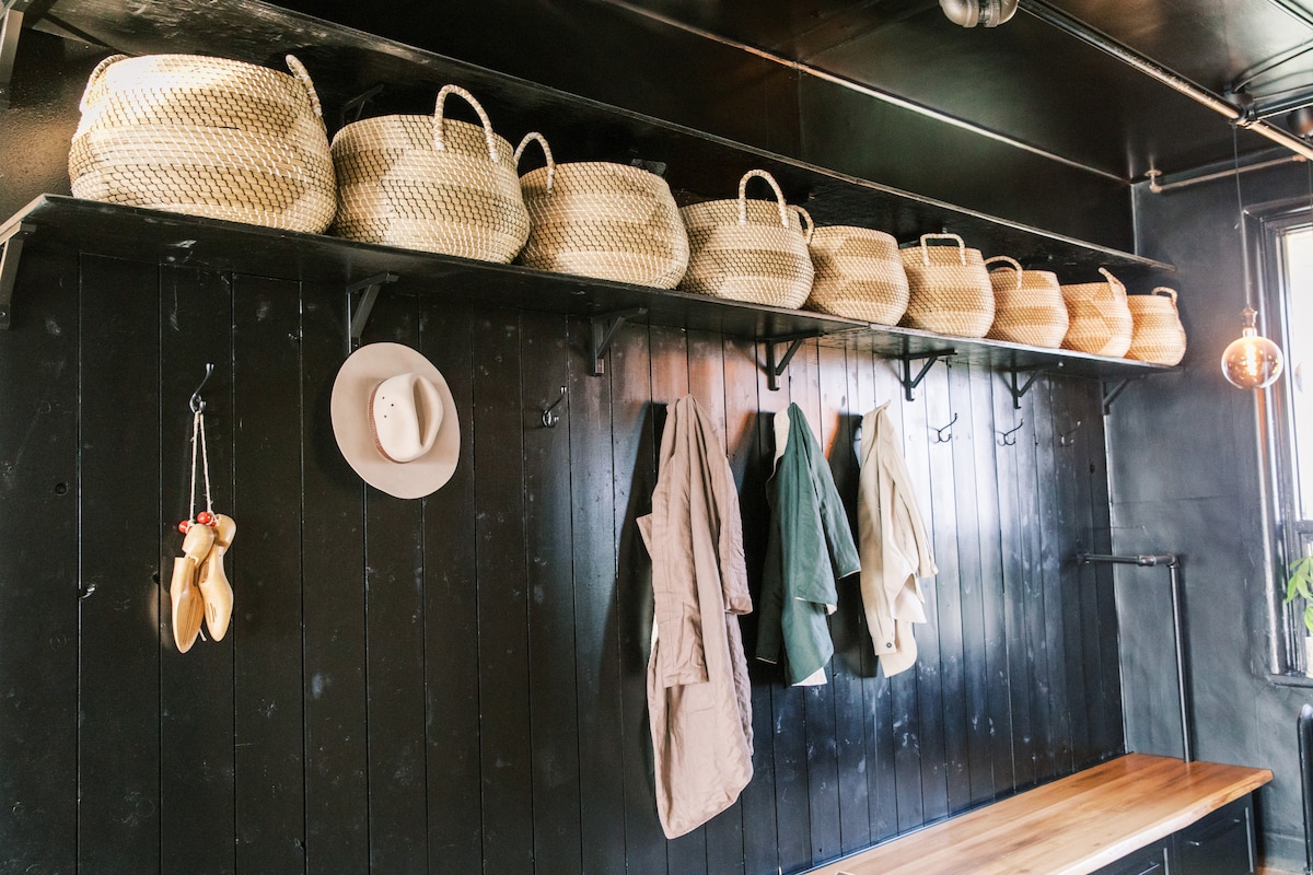 A wall featuring multiple woven baskets organized on a shelf, complemented by hooks with hanging jackets. A hat rests on one hook, and a wooden countertop offers additional space beneath the shelving.