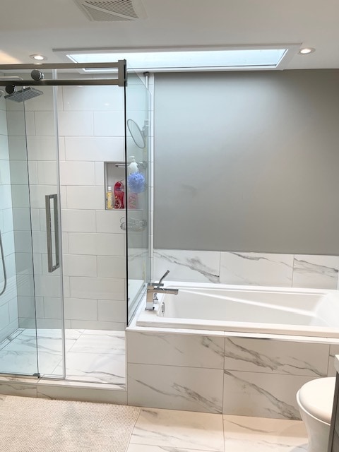 A modern bathroom is showcased, featuring a spacious glass shower enclosure with a rain showerhead, alongside a separate bathtub. Elegant marble tiles cover the walls and floor, while natural light enters through a skylight, enhancing the bright atmosphere.
