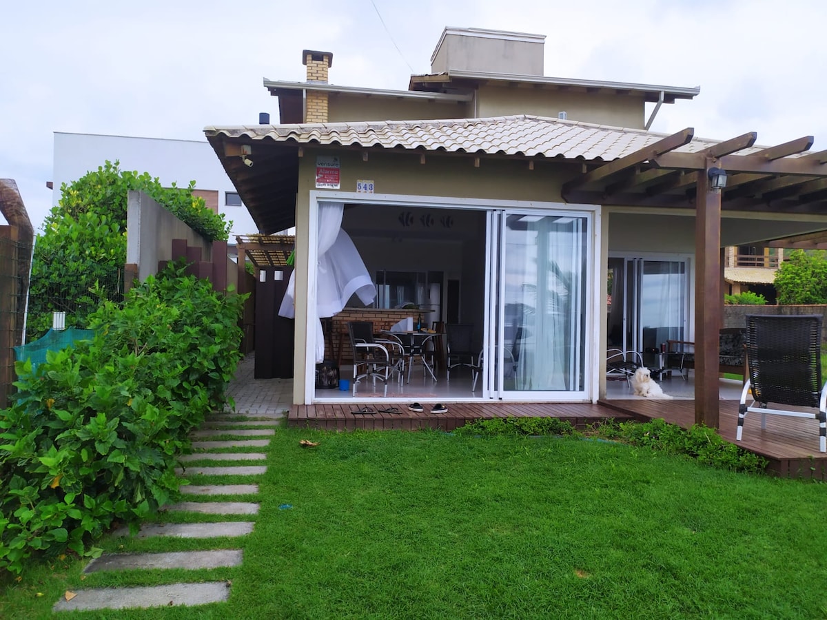 A spacious outdoor area is visible, featuring a neatly manicured lawn and paved stone pathway leading to a covered terrace. Glass doors open to an inviting dining space with a table and chairs, surrounded by greenery and partial ocean views. A gentle breeze flows through the area.