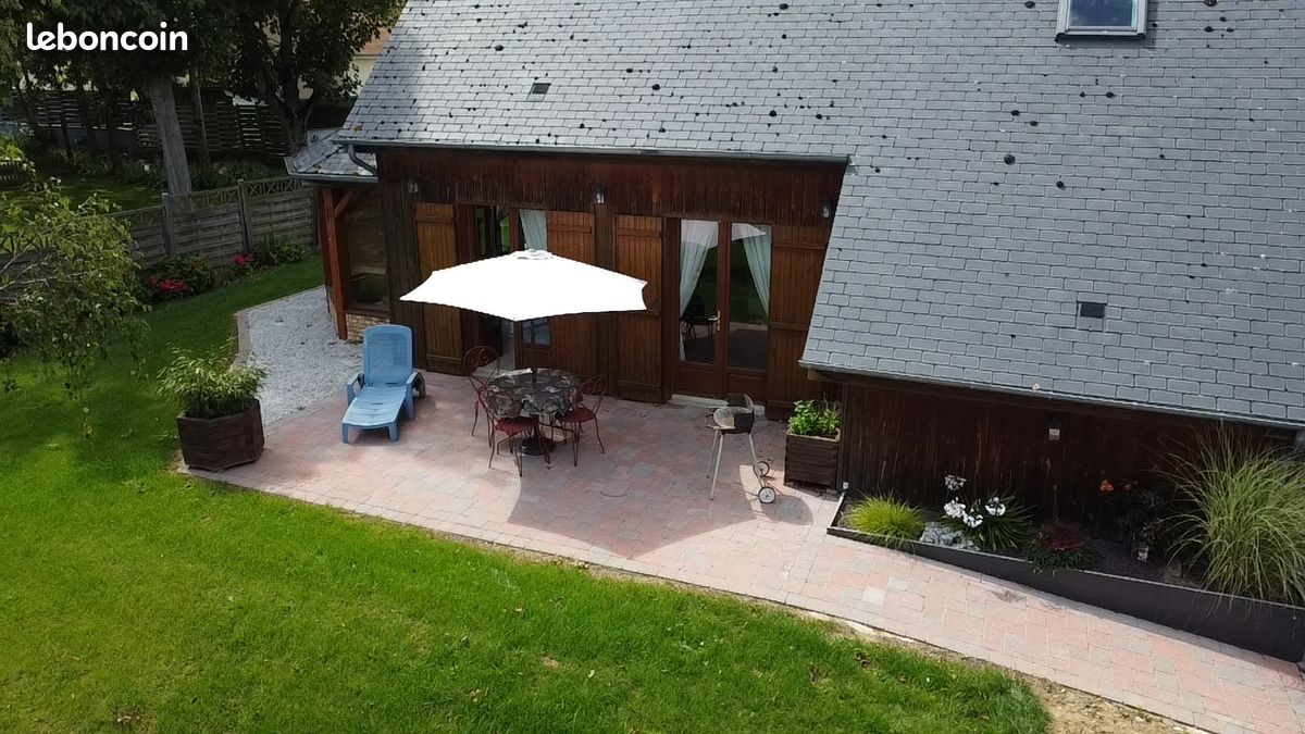 An outdoor seating area is depicted, featuring a table surrounded by chairs under a large umbrella. A lounge chair is positioned nearby on a paved patio, with a lush green lawn extending around the space. The wooden structure of the house is visible in the background.
