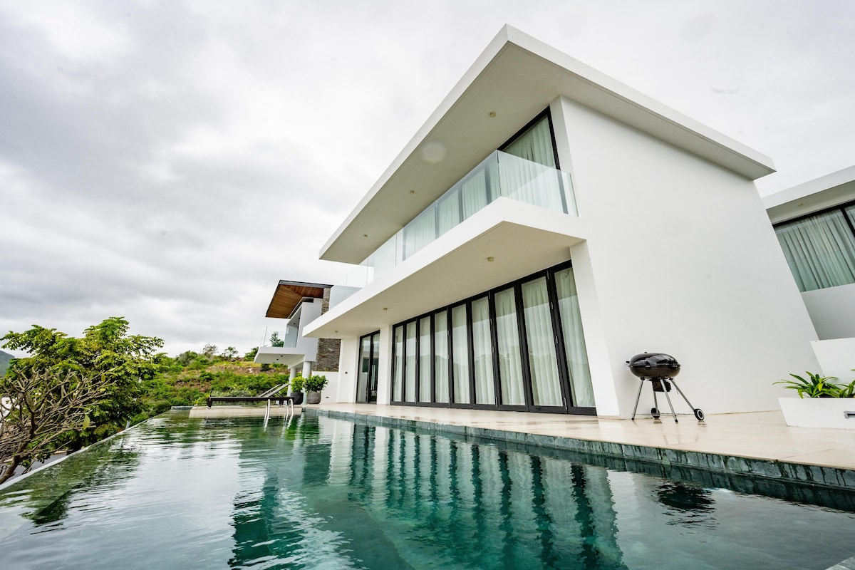 A modern villa is showcased with large glass doors reflecting the surrounding scenery. A swimming pool extends along the edge, enhancing the outdoor space. The structure features a clean white exterior, and the cloudy sky contrasts with the tranquil blue water.