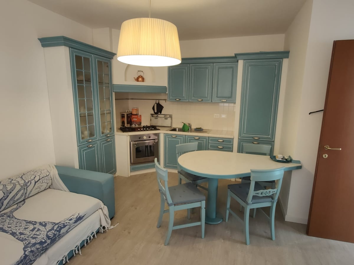 A kitchen area with teal cabinetry and a round dining table is highlighted. The space features a comfortable sofa covered with a light throw, an oven, and various kitchen appliances. Soft lighting is provided by a modern pendant light hanging above the dining table.