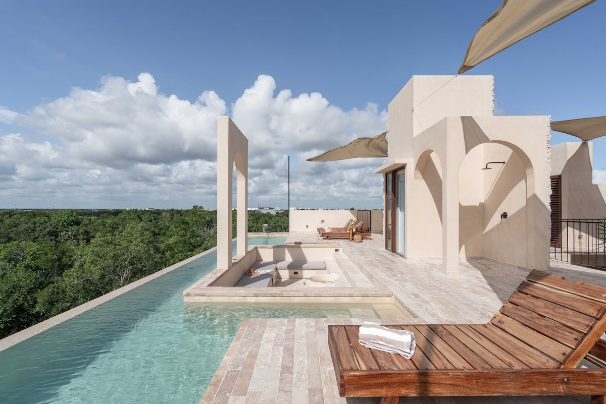 A private roof terrace features a sleek pool with a seamless edge blending into panoramic jungle views. Comfortable sun loungers are positioned alongside the water, beneath elegantly draped fabric awnings, providing shade. The terrace’s modern design is complemented by natural stone flooring.