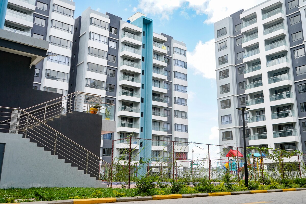 The image displays a modern multi-story apartment complex with a mix of grey and white facades. A staircase leads to the entrance, and a fenced outdoor play area is visible, featuring colorful playground equipment, surrounded by greenery and pathways.