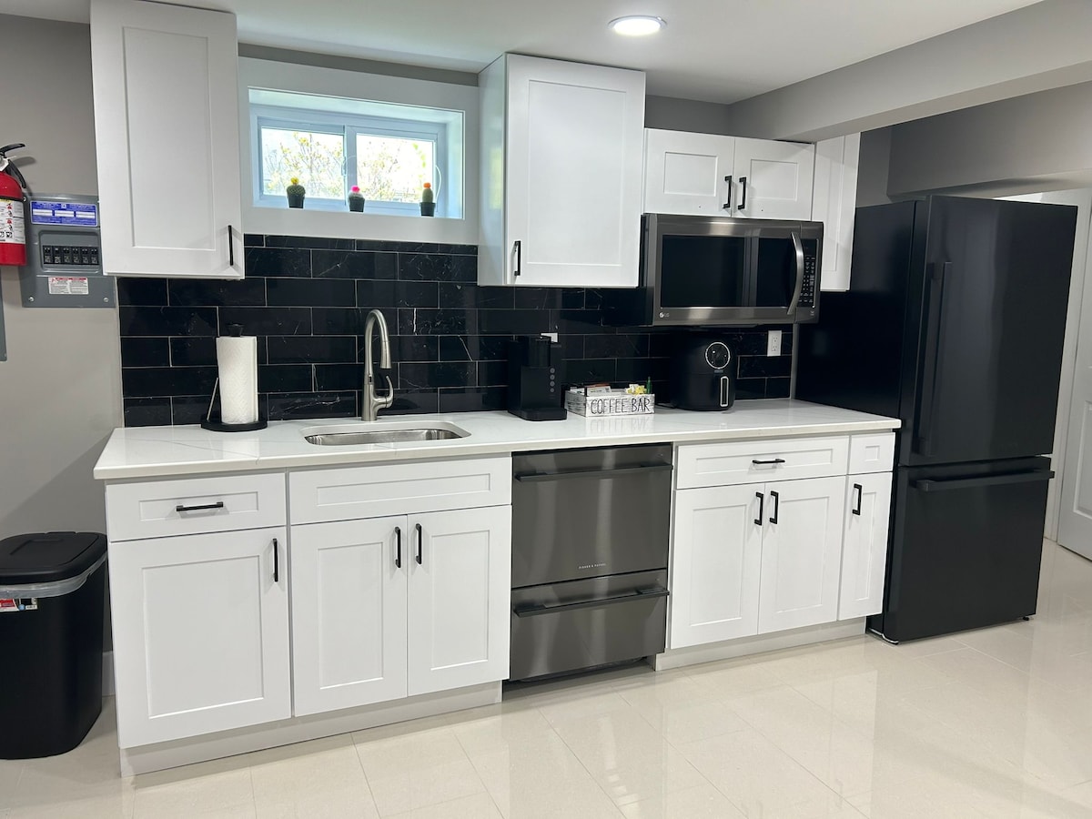 An open-plan kitchenette features modern white cabinetry, stainless steel appliances, and a black tiled backsplash. Natural light enters through a small window above the sink. A clean countertop provides ample space for cooking, while a black refrigerator and integrated oven are included.