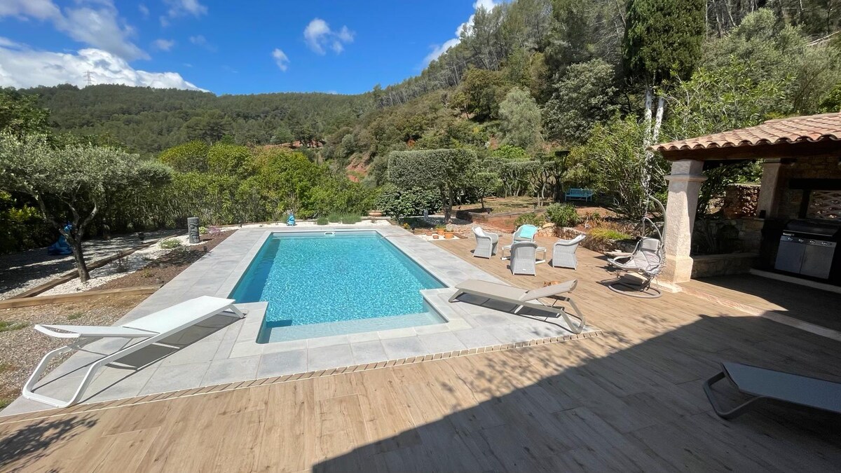 A spacious outdoor area features a well-maintained swimming pool surrounded by a sun terrace. Several lounge chairs are arranged around the pool, with views of the lush greenery and hills in the background. A shaded cooking area is also visible, enhancing the outdoor experience.