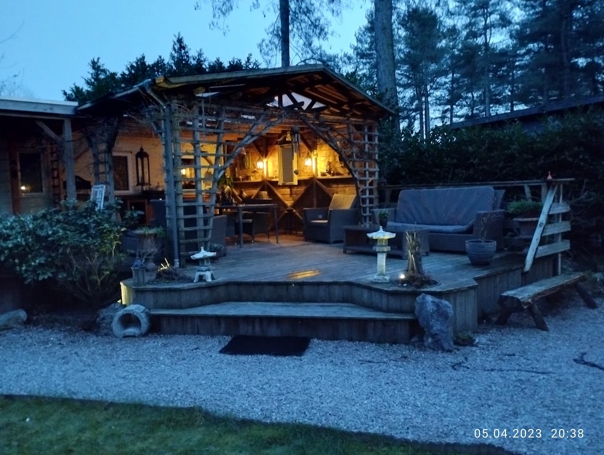An outdoor lounge area is presented under a wooden gazebo, surrounded by greenery. Comfortable seating is provided, with warm light illuminating the space, creating a cozy ambiance. Pathways of gravel lead to the area, inviting relaxation in this tranquil garden setting.