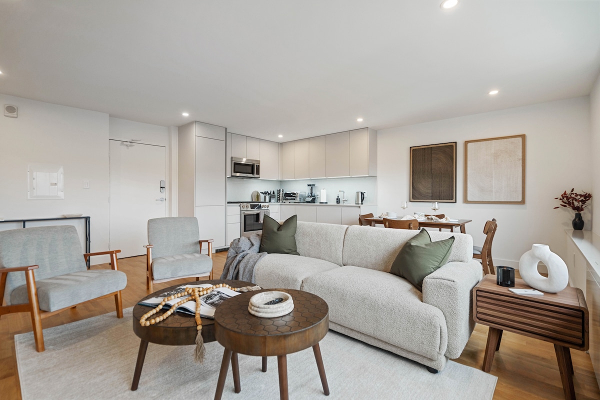 An open-concept living area showcases a comfortable sofa adorned with pillows, complemented by two accent chairs. A textured coffee table occupies the center, while a dining area with a table and chairs is visible in the background, alongside a modern kitchen equipped with stainless steel appliances.