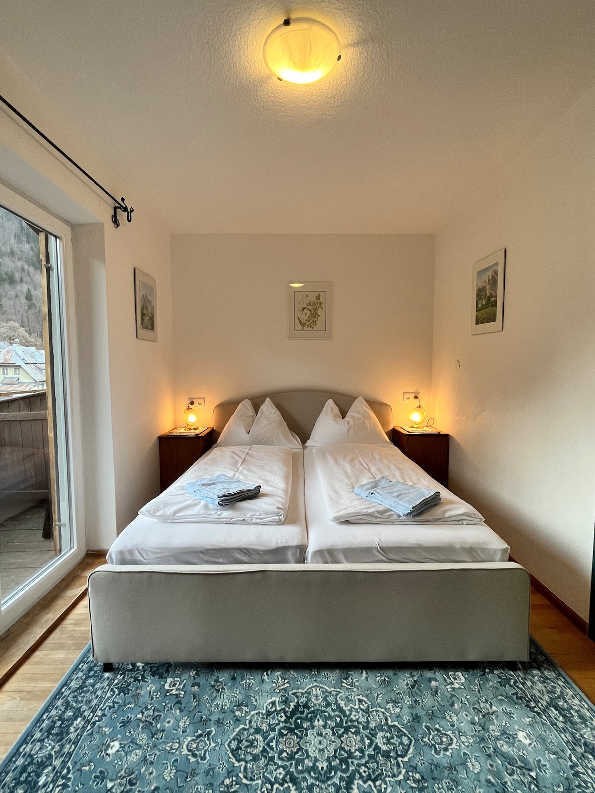 A bright bedroom features a spacious bed with crisp white linens and two matching decorative pillows. Soft lighting is provided by two bedside lamps. A balcony door allows natural light to enter the room, and a patterned rug adds warmth to the wooden floor.