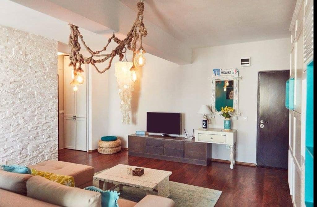 A cozy living area features a neutral sofa complemented by colorful cushions. A rustic coffee table is centered on a light rug. Unique lighting hangs from the ceiling, and a television is positioned against a wall. Decor items can be seen on a nearby shelf.