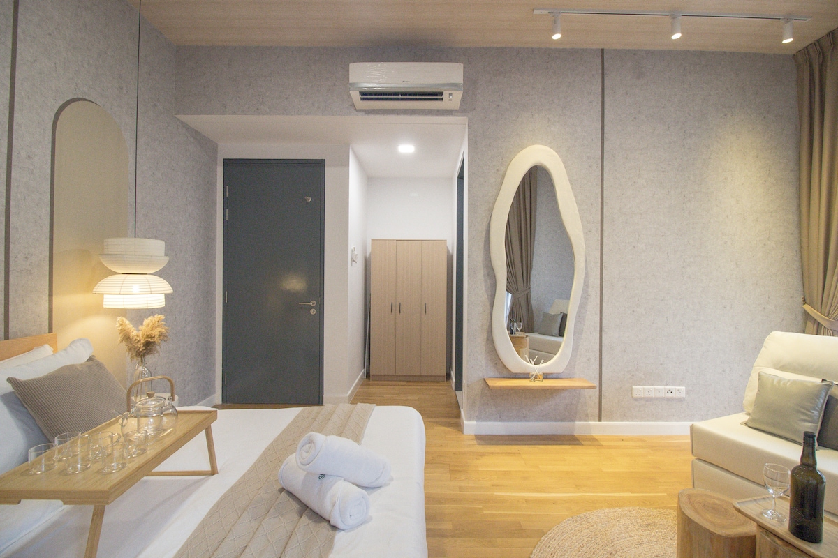 A serene bedroom features a plush bed with neatly arranged towels and a tray presenting glasses. A light wood accent wall complements the modern furnishings, including a plush sofa and a uniquely shaped mirror. Soft natural light filters in, enhancing the tranquil ambiance.