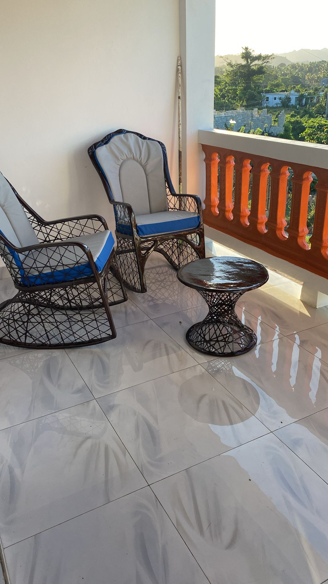 A small outdoor balcony features two wicker chairs with blue cushions and a round glass table. The tiled floor reflects sunlight, creating a bright ambiance. A wooden railing adds a warm touch, and distant greenery is visible beyond the balcony.