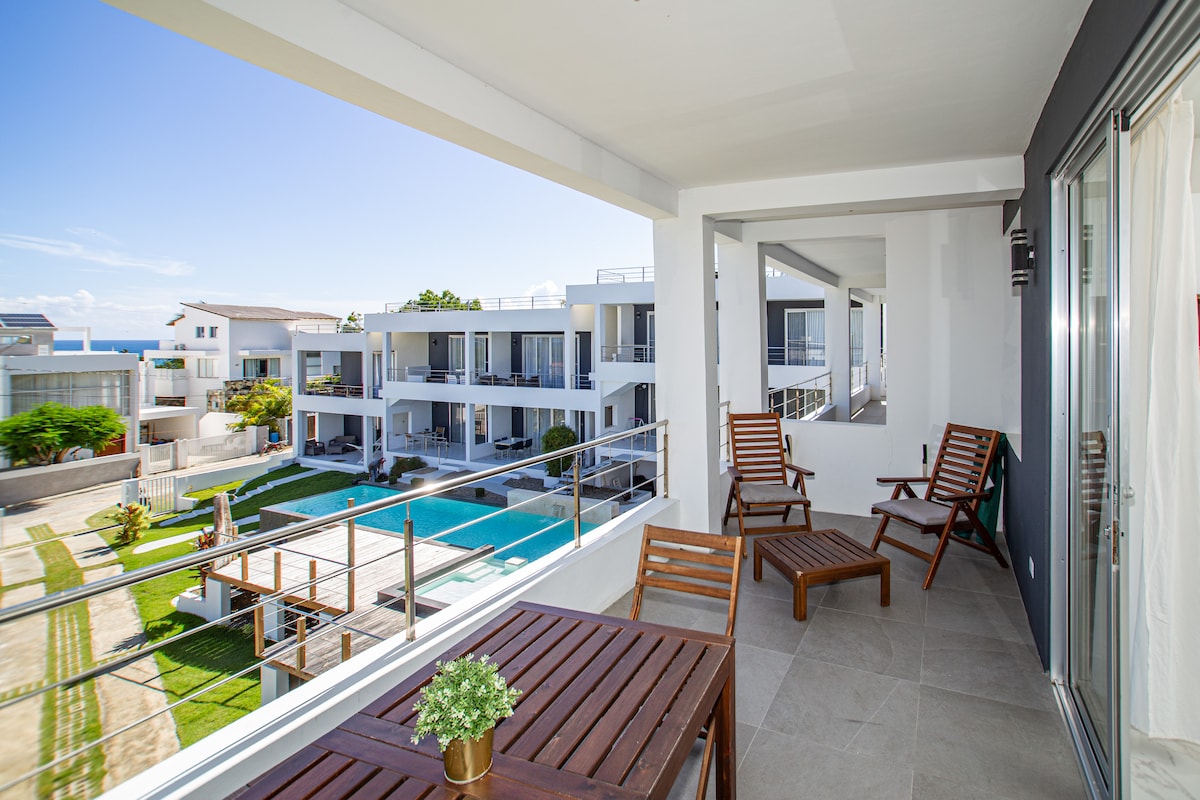 A private balcony features a wooden table and two chairs, inviting relaxation. The view includes a landscaped garden and a large infinity swimming pool surrounded by modern structures. Natural light illuminates the space, with ocean glimpses visible in the background.