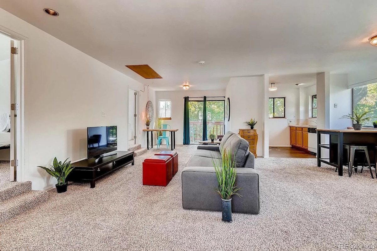 An open living space is presented with soft carpet underfoot, featuring a comfortable sofa and a distinct coffee table. Large windows allow natural light to illuminate the area. A dining table is nearby, complemented by a kitchen view in the background.